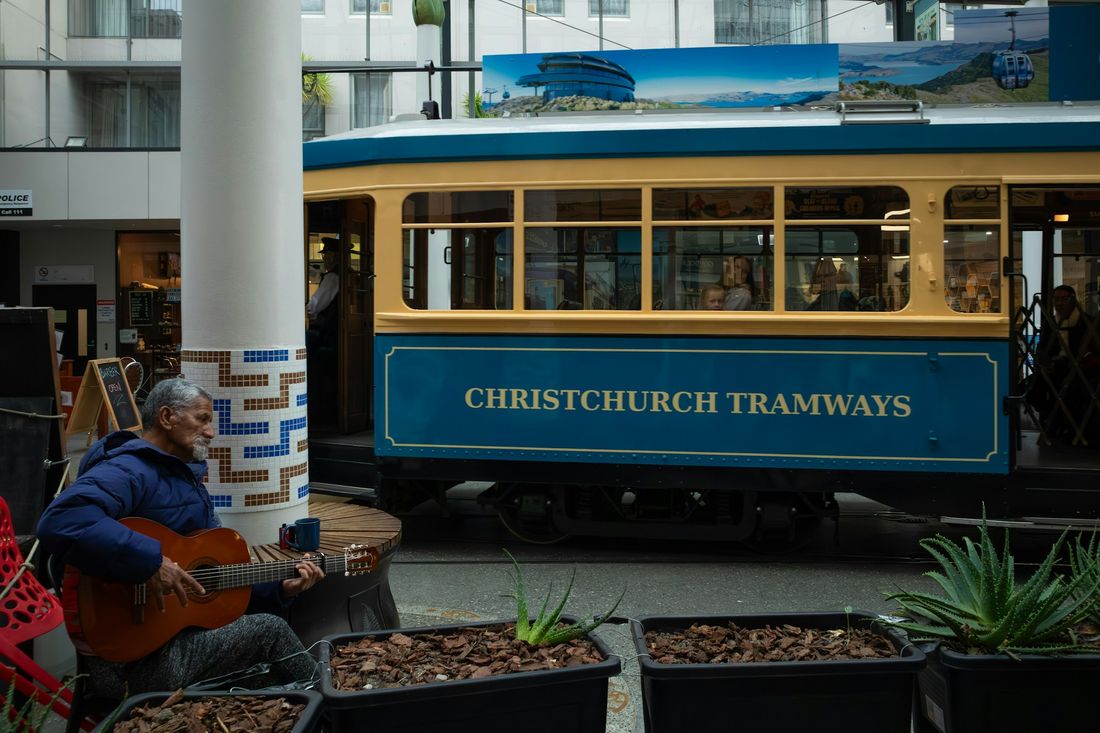 A street musician playing acoustic guitar beside a blue and yellow Christchurch tram