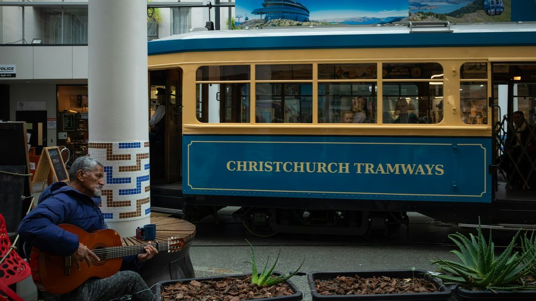 A street musician playing acoustic guitar beside a blue and yellow Christchurch tram