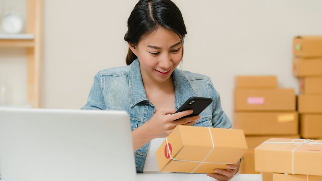 An online seller scanning a parcel with her phone next to a laptop and stacked boxes