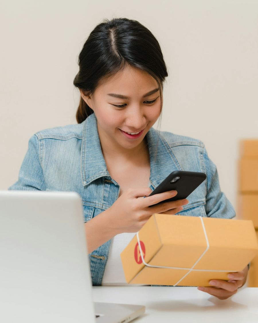 An online seller scanning a parcel with her phone next to a laptop and stacked boxes