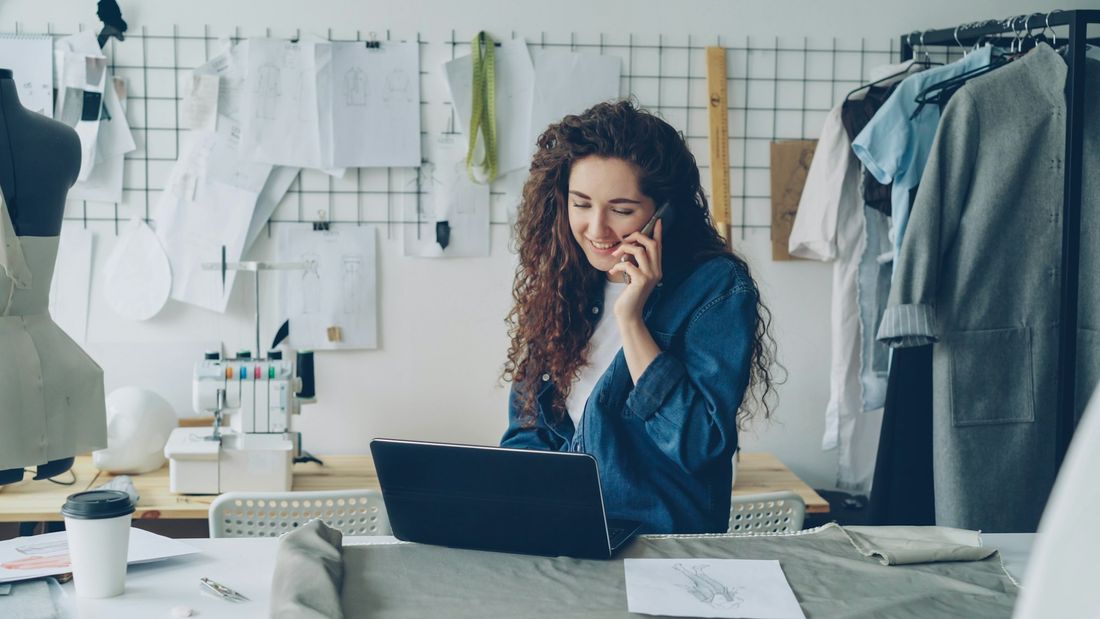 A fashion start-up founder on the phone at her laptop with a sewing machine and garments behind her