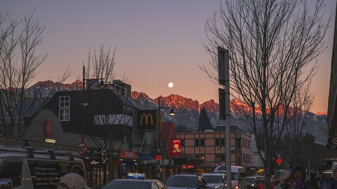 Shops and restaurants on a Queenstown street below snow-capped mountains at dusk